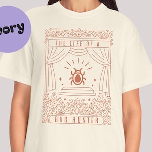 Puede incluir: Camiseta color marfil con un gráfico marrón de un insecto en un escenario con cortinas y el texto "THE LIFE OF A BUG HUNTER". La camiseta tiene cuello redondo y mangas cortas.