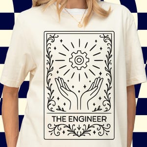 May include: A cream-coloured t-shirt with a black and white graphic. The design features a gear radiating light, held by two hands, with the text "THE ENGINEER" below. The graphic is framed with decorative flourishes.