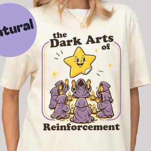 Op de afbeelding: Crèmekleurig T-shirt met een cartoonafbeelding. De afbeelding toont een lachende gele ster, figuren in paarse gewaden en de tekst "the Dark Arts of Reinforcement". Een paarse cirkel met "Natural" staat linksboven.