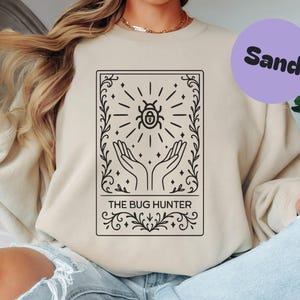 May include: A sand-coloured sweatshirt with a black graphic of a beetle, radiating lines, hands, and the text "THE BUG HUNTER." A purple circle with the word "Sand" is in the upper right corner. The sweatshirt is a neutral colour.
