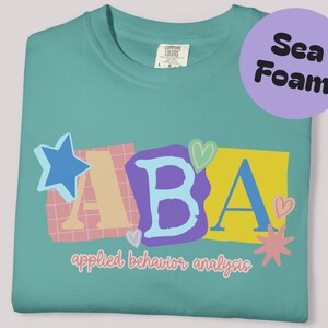 Könnte beinhalten: Meergrünes T-Shirt mit den Buchstaben "ABA" in einem farbenfrohen, verspielten Design. Die Buchstaben sind vor einem Stern, Herzen und geometrischen Formen platziert. Darunter steht der Schriftzug "applied behavior analysis".