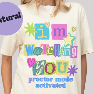 May include: Cream-colored t-shirt with the text "I'm Watching You" in colorful, blocky letters and a purple circle that says "Natural". The phrase "proctor mode activated" is below the main text. The design includes hearts and stars.