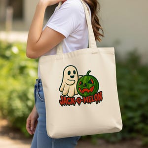 May include: Beige tote bag featuring a cartoon ghost and a watermelon carved like a jack-o'-lantern. The text "JACK-O-MELON" is printed in red and black below the images. The bag is held by a person.
