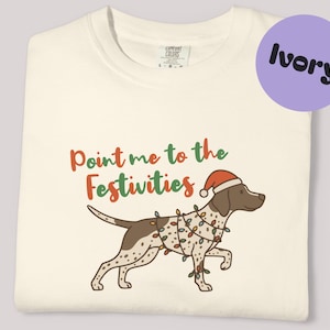 May include: Ivory t-shirt with a festive design. The graphic features a brown and white dog wearing a Santa hat and Christmas lights, with the text "Point me to the Festivities" in red and green.