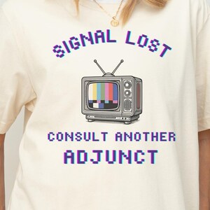 Puede incluir: Camiseta color crema con un gráfico de televisión retro que muestra una pantalla de prueba. El texto encima del televisor dice "SIGNAL LOST" y debajo, "CONSULT ANOTHER ADJUNCT", todo en fuente pixelada.