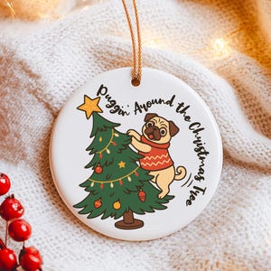 May include: A white ceramic Christmas ornament with a pug dog wearing a red sweater, decorating a green Christmas tree. The ornament has the text "Puggin' Around the Christmas Tree" in black script and a gold string for hanging.