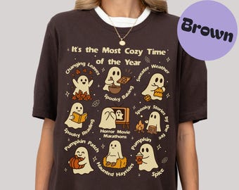 Könnte beinhalten: Braunes T-Shirt mit weißem Kragen. Die Grafik zeigt Geister und Text, darunter "It's the Most Cozy Time of the Year" und Illustrationen von Herbstaktivitäten wie Backen und Lesen.
