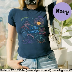 May include: A navy blue short-sleeve t-shirt featuring a colourful graphic print with the text 'tis the season' surrounded by illustrations of a campfire, a sunset, a monster ice cream cone, a tent, and a ghost in a floatie with Halloween motifs.
