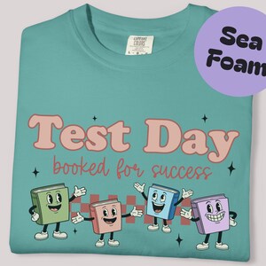 Könnte beinhalten: Meergrünes T-Shirt mit dem Text "Test Day booked for success" in Rosa. Das Shirt zeigt Cartoon-Bücher mit Armen und Beinen. Ein lila Kreis mit den Worten "Sea Foam" befindet sich in der oberen rechten Ecke.