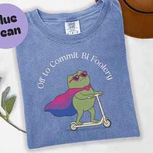 May include: Blue jean t-shirt featuring a green frog wearing heart-shaped sunglasses and a cape in the bisexual pride flag colours. The text "Off to Commit Bi Foolery" is printed on the shirt.