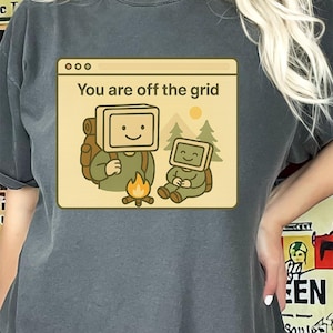 May include: Dark gray t-shirt featuring a graphic of two cartoon computer-headed figures camping. The graphic includes the text "You are off the grid" in a window-like frame, with a campfire, trees, and a sun.