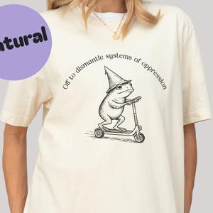 Puede incluir: Camiseta color crema con un gráfico de una rana con sombrero de mago en un patinete. El texto "Off to dismantle systems of oppression" está sobre la rana. Un círculo morado con "Natural" está en la parte superior izquierda.