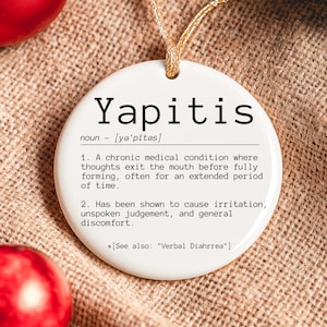 Op de afbeelding: Een witte, ronde ornament met het woord "Yapitis" in zwarte letters en de definitie. Het hangt aan een gouden draad tegen een getextureerde, bruine achtergrond. Twee rode ornamenten zijn ook zichtbaar.