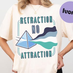 May include: An ivory t-shirt featuring a graphic of a blue prism refracting light into a wavy banner with dark blue, light blue, white, and teal stripes. The text "REFRACTION NO ATTRACTION" is printed in teal and blue letters above and below the prism.