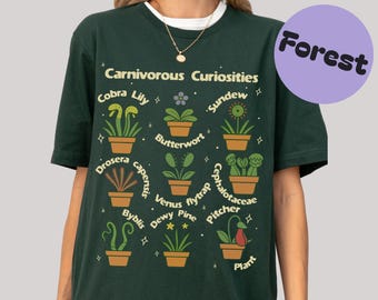 Può includere: T-shirt verde scuro con la scritta "Carnivorous Curiosities" e illustrazioni di varie piante carnivore, tra cui Cobra Lily e Sundew. Un cerchio viola con la parola "Forest" si trova nell'angolo in alto a destra.