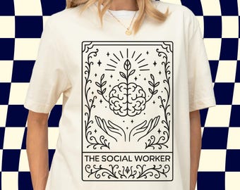 Puede incluir: Una camiseta de color crema con un gráfico de líneas negras de un cerebro con una planta brotando sobre dos manos abiertas, enmarcado por adornos. El texto THE SOCIAL WORKER está impreso en un recuadro rectangular en la parte inferior.