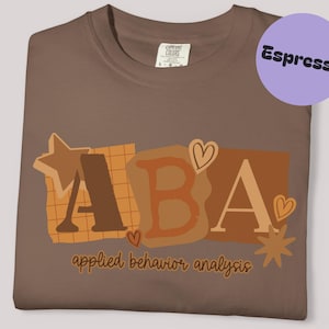 以下が含まれることがあります： エスプレッソ色のTシャツ。コラージュデザインで「ABA」の文字が描かれています。デザインには星、ハート、「applied behavior analysis」の文字が含まれています。右上には「Espresso」という文字が入った紫色の円があります。