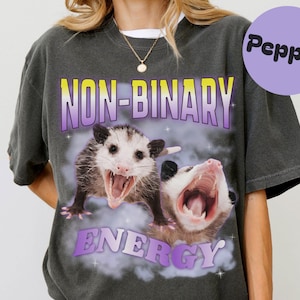 Könnte beinhalten: Dunkelgraues T-Shirt mit den Worten "NON-BINARY ENERGY" in Gelb und Lila, mit zwei Opossums mit offenem Mund. Ein lila Kreis mit dem Wort "Pepper" befindet sich in der oberen rechten Ecke.