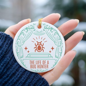 May include: A white ceramic ornament with a gold hanging loop. The ornament features a teal and orange bug design with the text "THE LIFE OF A BUG HUNTER". The design includes a stage with curtains and stars.