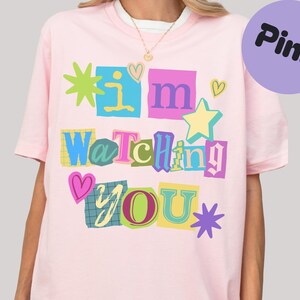 Könnte beinhalten: Hellrosa T-Shirt mit dem Schriftzug "I'm Watching You" in einem farbenfrohen Collage-Design. Das Design umfasst Sterne, Herzen und das Wort "Pink" in einem lila Kreis.