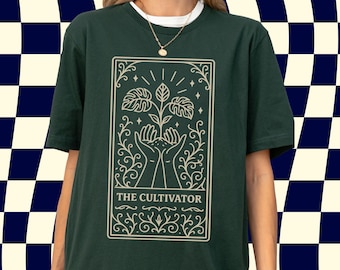 Puede incluir: Camiseta verde oscuro con un gráfico color crema de una planta que crece de manos, enmarcada por elementos decorativos. El texto "THE CULTIVATOR" está en la parte inferior. La camiseta se usa sobre una camiseta interior blanca y un collar dorado.