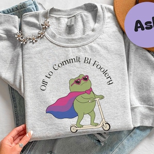 May include: Light gray sweatshirt with the text "Off to Commit Bi Foolery" around a cartoon frog wearing sunglasses and a cape in the colors of the bisexual pride flag. The frog is riding a scooter.