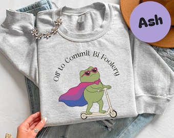 Può includere: Felpa grigio chiaro con la scritta "Off to Commit Bi Foolery" attorno a un ranocchio dei cartoni animati con occhiali da sole e un mantello con i colori della bandiera del pride bisessuale. Il ranocchio va in scooter.