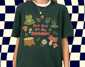 May include: Dark green t-shirt with a whimsical design featuring a frog, mushrooms, and the text "Get Out Of My Swamp!". The design includes a tree stump, berries, and a daisy. The shirt has short sleeves and a round neckline.