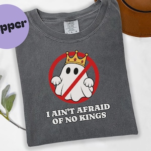 Könnte beinhalten: Ein anthrazitfarbenes T-Shirt mit einer Grafik eines weißen Geistes, der eine goldene Krone trägt und sich in einem roten Kreis mit einem diagonalen Strich befindet. Unter der Grafik steht der Text I AIN'T AFRAID OF NO KINGS in weißen Großbuchstaben.