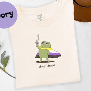 May include: Ivory-colored t-shirt featuring a cartoon frog wearing sunglasses, a cape with non-binary colors, and holding a sword. The text "they/them" is printed below the frog. The shirt has a Comfort Colors tag.