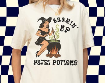 May include: Off-white t-shirt with a vintage-style graphic of a witch stirring a cauldron over a fire. The words "BREWIN' UP" and "PETRI POTIONS" are printed above and below the image. The witch is in a black dress and hat.