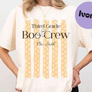 May include: An ivory t-shirt featuring a graphic with five vertical orange polka-dot stripes. The text reads Third Grade Boo Crew with two small ghost illustrations and the name Mrs. Smith printed in a script font below the main text.