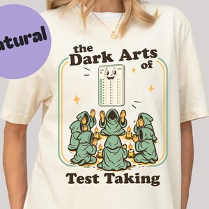 Könnte beinhalten: Cremefarbenes T-Shirt mit dem Text "the Dark Arts of Test Taking" und einer Grafik eines Testformulars mit Gesicht, umgeben von Kapuzenfiguren und Kerzen. Ein violetter Kreis mit "Natural" befindet sich oben links.