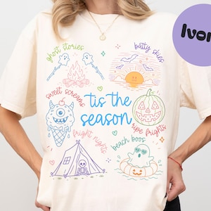 May include: An ivory t-shirt featuring a colorful graphic print with the text 'tis the season' in the center. The design includes illustrations of a campfire, ghosts, a sunset with bats, a monster ice cream cone, a jack-o'-lantern, and a tent.