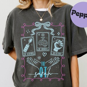 Puede incluir: Una camiseta gris carbón de manga corta con un diseño gráfico en azul claro y rosa. El estampado muestra tres cartas estilo tarot etiquetadas como The Pathfinder con una cuchara, The Supporter con un cerebro y The Restorer con un puño cerrado.