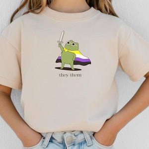 May include: Beige t-shirt featuring a cartoon frog wearing sunglasses, a cape in non-binary colors, and holding a sword. The text "they/them" is printed below the frog. The shirt is made of a soft material.