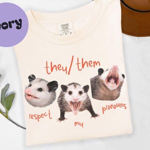 Peut inclure: T-shirt ivoire avec un motif de trois opossums et le texte "they/them pronouns". Les mots "respect my" sont également imprimés sur le t-shirt. Un cercle violet avec le mot "ivory" est dans le coin supérieur gauche.