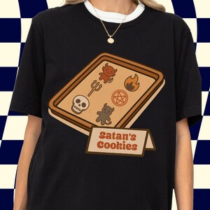 May include: Black t-shirt featuring a graphic of a tray of cookies with devilish symbols and the text "Satan's Cookies." The cookies include a skull, a trident, a flame, a pentagram, and devil figures. The shirt has a white collar.