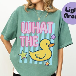 Peut inclure: Un t-shirt à manches courtes vert clair avec un imprimé graphique comportant le texte WHAT THE en lettres roses style bulle au-dessus d'un canard en caoutchouc jaune. Le motif inclut des rayures verticales bleu clair et trois étoiles colorées.
