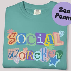 May include: Seafoam green t-shirt with the words "SOCIAL WORKER" in a colorful, cut-out style design. The design includes hearts, stars, and other decorative elements. The shirt is folded neatly.