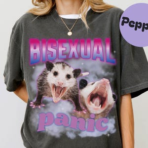 May include: Dark gray t-shirt with the words "BISEXUAL panic" in pink and blue, featuring two opossums with open mouths. A purple circle with the word "Pepper" is in the upper right corner.
