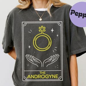 May include: Dark gray t-shirt featuring a tarot card design with a yellow and white graphic. The graphic includes a symbol, hands, and the text "THE ANDROGYNE". A purple circle with the word "Pepper" is in the upper right corner.