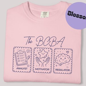 Op de afbeelding: Een lichtroze t-shirt met de tekst "The BCBA" in donkerblauwe letters. Daaronder drie geïllustreerde vakken met de woorden "Analyst", "Motivator" en "Regulator". Een paarse cirkel met het woord "Blossom" staat rechtsboven.