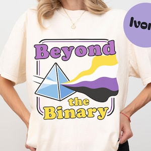 May include: Ivory t-shirt with a graphic design. The design features the words "Beyond the Binary" in purple and yellow, a prism, and a non-binary flag. A purple circle with the word "ivory" is in the upper right corner.
