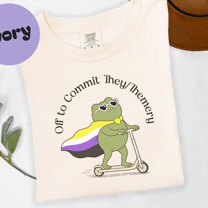 May include: Ivory t-shirt featuring a cartoon frog wearing sunglasses and a non-binary pride flag cape, riding a scooter. The text around the frog reads "Off to Commit They/Themery."