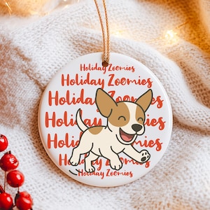 May include: A white ceramic holiday ornament with a tan and white cartoon dog and the words "Holiday Zoomies" in red. The ornament has a gold string for hanging and is set against a white knitted background.
