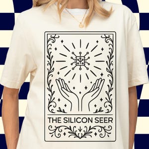 May include: A cream-coloured t-shirt with a black and white graphic. The design shows a circuit board with radiating lines, hands reaching upwards, and the text "THE SILICON SEER" within a decorative frame. This is a casual wear item.