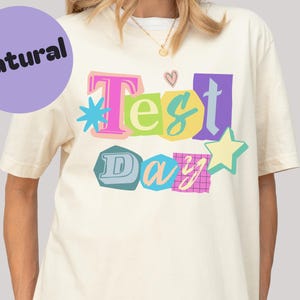 May include: Cream-colored t-shirt with the text "Test Day" in colorful, cut-out style letters. The design includes a star, a heart, and other decorative elements. A purple circle with the word "Natural" is in the upper left corner.