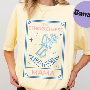 Könnte beinhalten: Hellgelbes T-Shirt mit einem blau-rosa Grafikdesign. Der Aufdruck zeigt "THE STRING CHEESE MAMA" mit einer Illustration von Käsefäden und Händen. Ein violetter Kreis mit "Banana" befindet sich oben rechts.