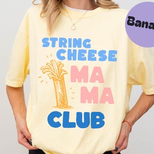 Könnte beinhalten: Hellgelbes T-Shirt mit den Worten "STRING CHEESE MAMA CLUB" in Blau und Rosa. Eine Cartoon-Illustration von Käsefäden befindet sich in der Mitte. Ein lila Kreis mit dem Wort "Banana" befindet sich in der oberen rechten Ecke.